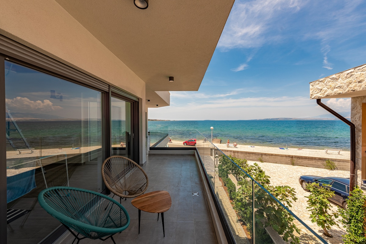 A modern balcony features two stylish chairs and a small table, overlooking a sandy beach and calm sea. The expansive glass panels offer unobstructed views of the water and sky, creating a seamless connection to the outdoor environment.