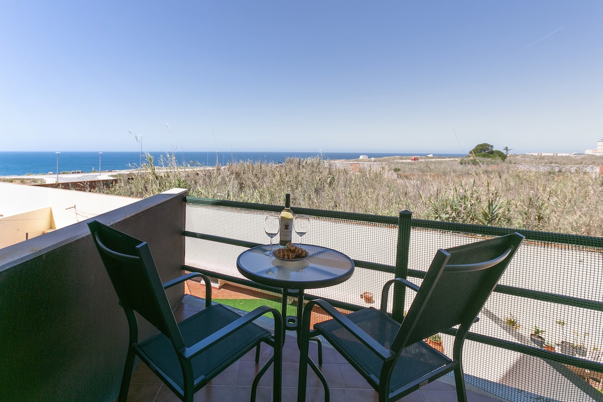 A balcony offers a round table with two chairs and a scenic view of the ocean. A bottle of wine and two glasses are displayed on the table, with the horizon visible in the distance and greenery framing the space.