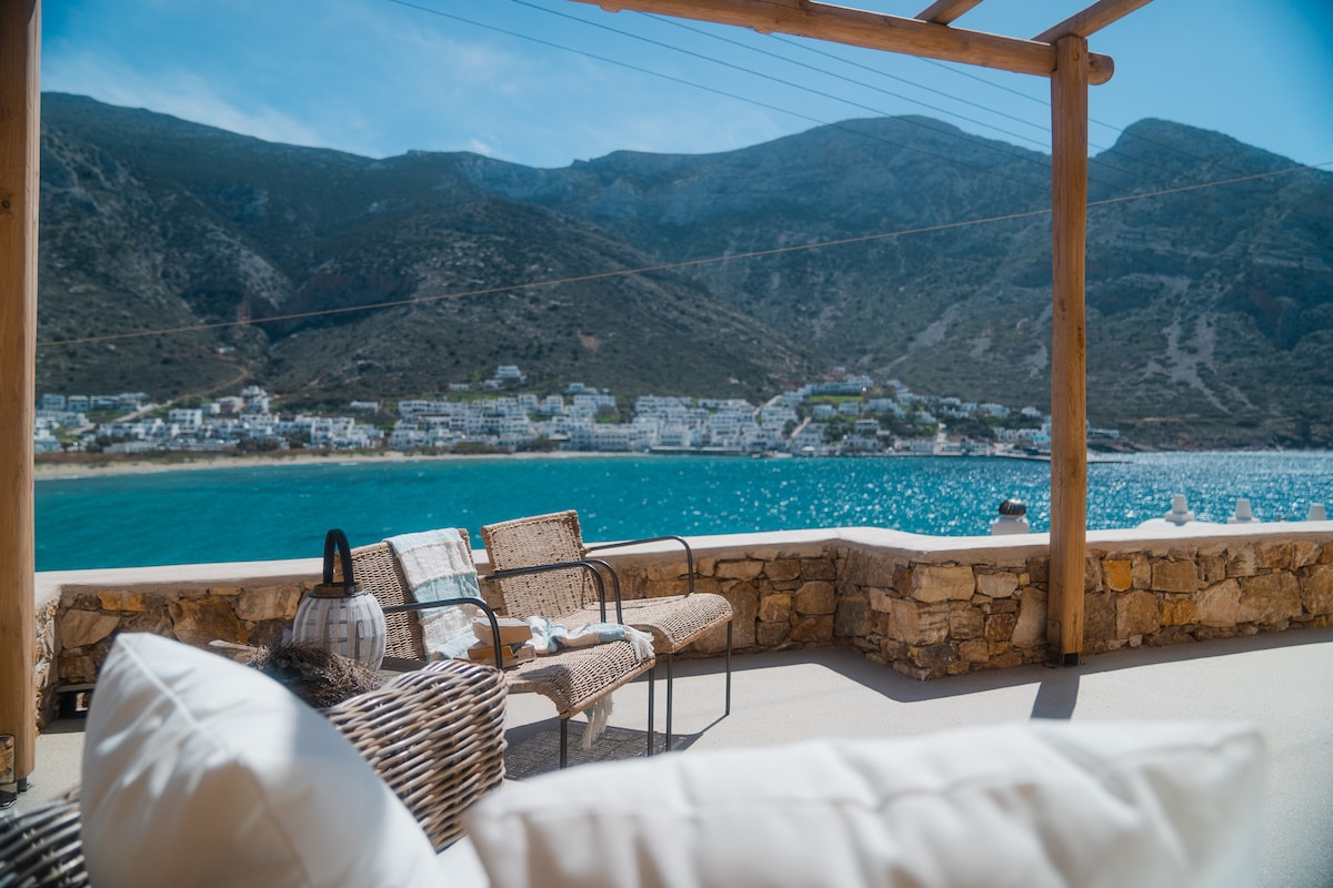 A spacious balcony offers a view of the shimmering blue sea and coastal mountains. Comfortable seating is arranged under a wooden pergola, providing a relaxing space to enjoy the scenery. The surrounding stone wall adds a natural touch to the setting.