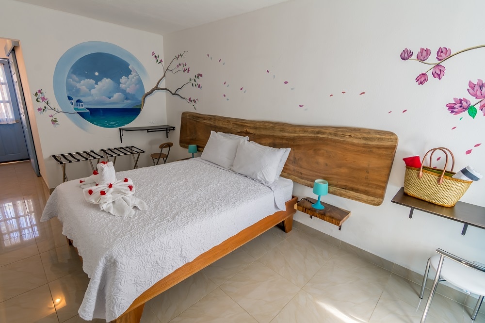 A bright sleeping area features a spacious bed with a textured white coverlet. Soft blue lamps are positioned on each side atop wooden shelves. A vibrant mural depicting clouds and flowers adds an artistic touch, while a basket and a piece of furniture complete the room's functionality.