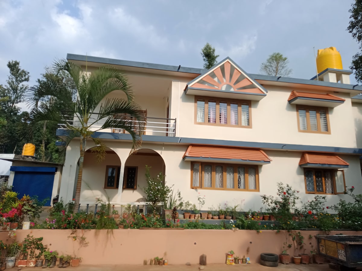 A two-story house is presented with a charming facade, featuring a combination of sloped roofs and decorative elements. Lush greenery surrounds the building, with a variety of flowering plants in pots adding vibrant colors along the foundation. A water tank is visible on the roof.