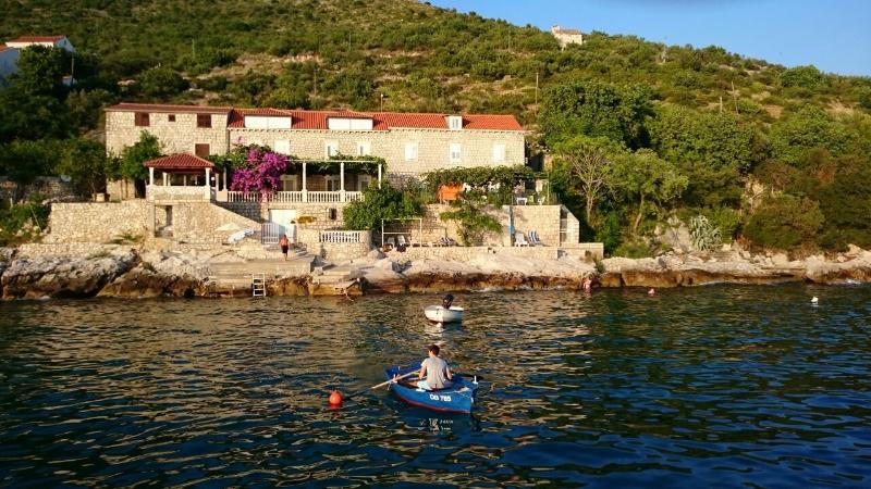 A quaint stone property is nestled against a lush hillside, surrounded by Mediterranean greenery. Gentle waves lap against the rocky shore, where individuals can be seen in small boats, creating a serene scene. The vibrant purple flowers and terraced areas add to the charm of the setting.