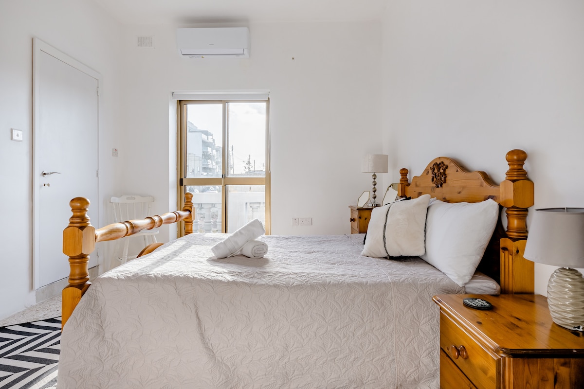 A well-lit private room features a spacious double bed adorned with neutral bedding. A window allows natural light to fill the space. Accompanying wooden furniture includes a nightstand and a lamp, creating a calm environment. An air conditioning unit is positioned above the bed.