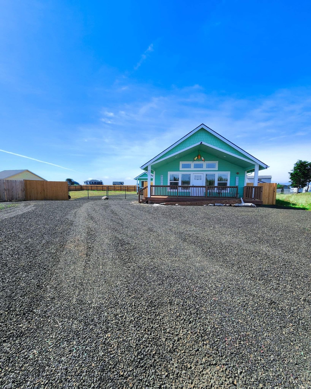 A charming house with a green exterior is set against a clear blue sky. A spacious gravel driveway leads to the front, with a covered porch visible. Wooden fencing encloses the property, providing a sense of privacy. The surrounding landscape features open space.