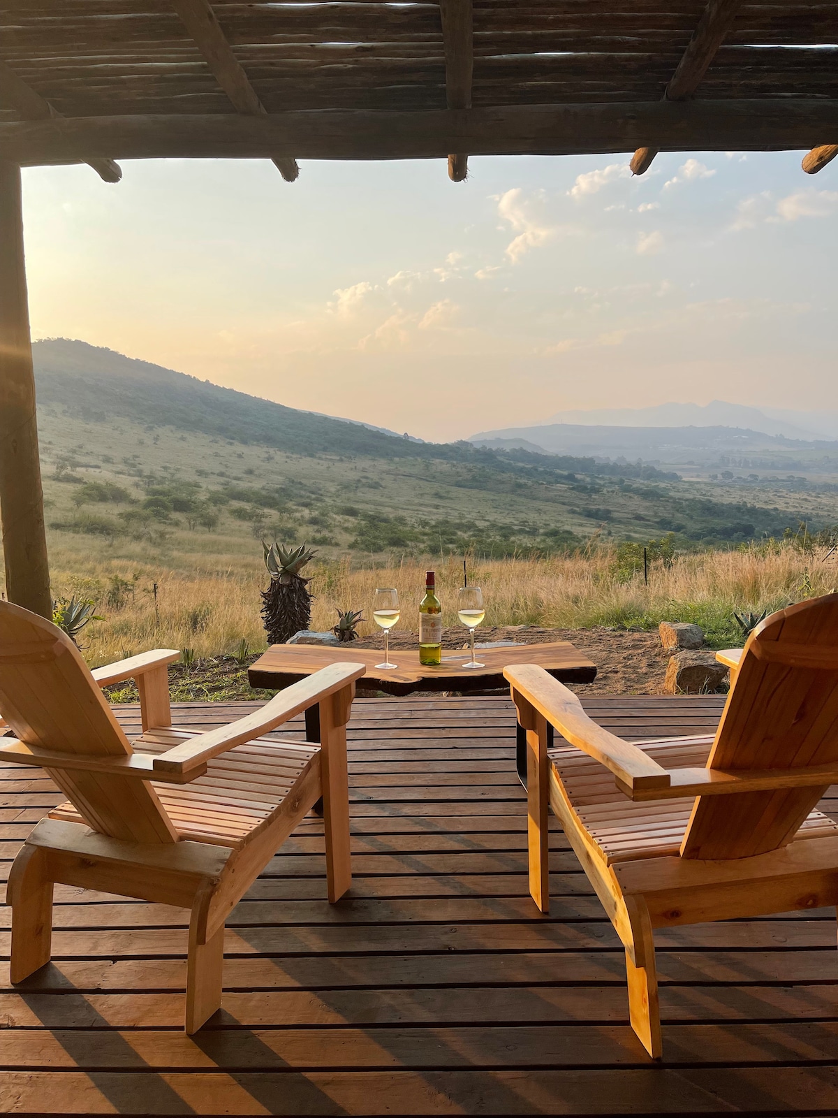 Two wooden chairs are positioned on a deck, facing a scenic landscape of rolling hills and distant mountains. A small table holds a bottle of wine and two glasses, set against the backdrop of a soft, hazy sky.