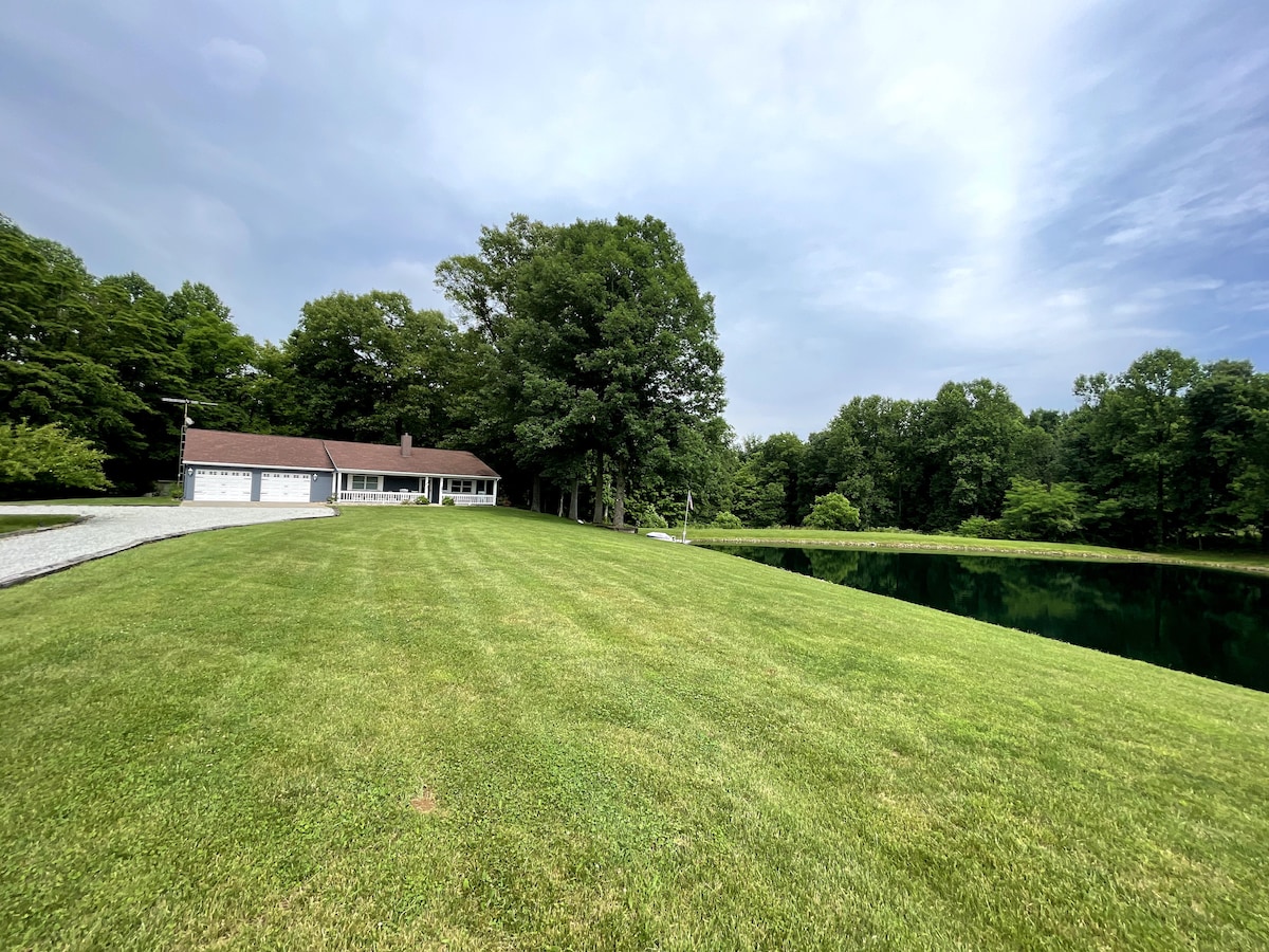 The image shows a well-kept lawn leading to a tranquil, reflective pond. A simple single-story house is positioned to the left, with a driveway visible. Lush green trees surround the area, creating a serene atmosphere.