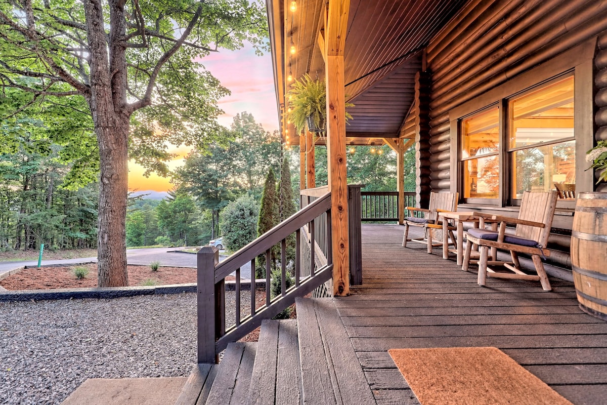 The front porch features a welcoming entry with wooden railings and rocking chairs, surrounded by trees. The view showcases the serene landscape at sunset, with hues of orange and pink illuminating the sky, enhancing the natural beauty of the surroundings.