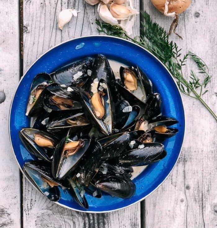A blue plate holds an assortment of freshly cooked mussels, glistening in a light broth. Surrounding the plate are a few cloves of garlic and a sprig of fresh herbs, enhancing the presentation. The rustic wooden surface provides a natural backdrop.