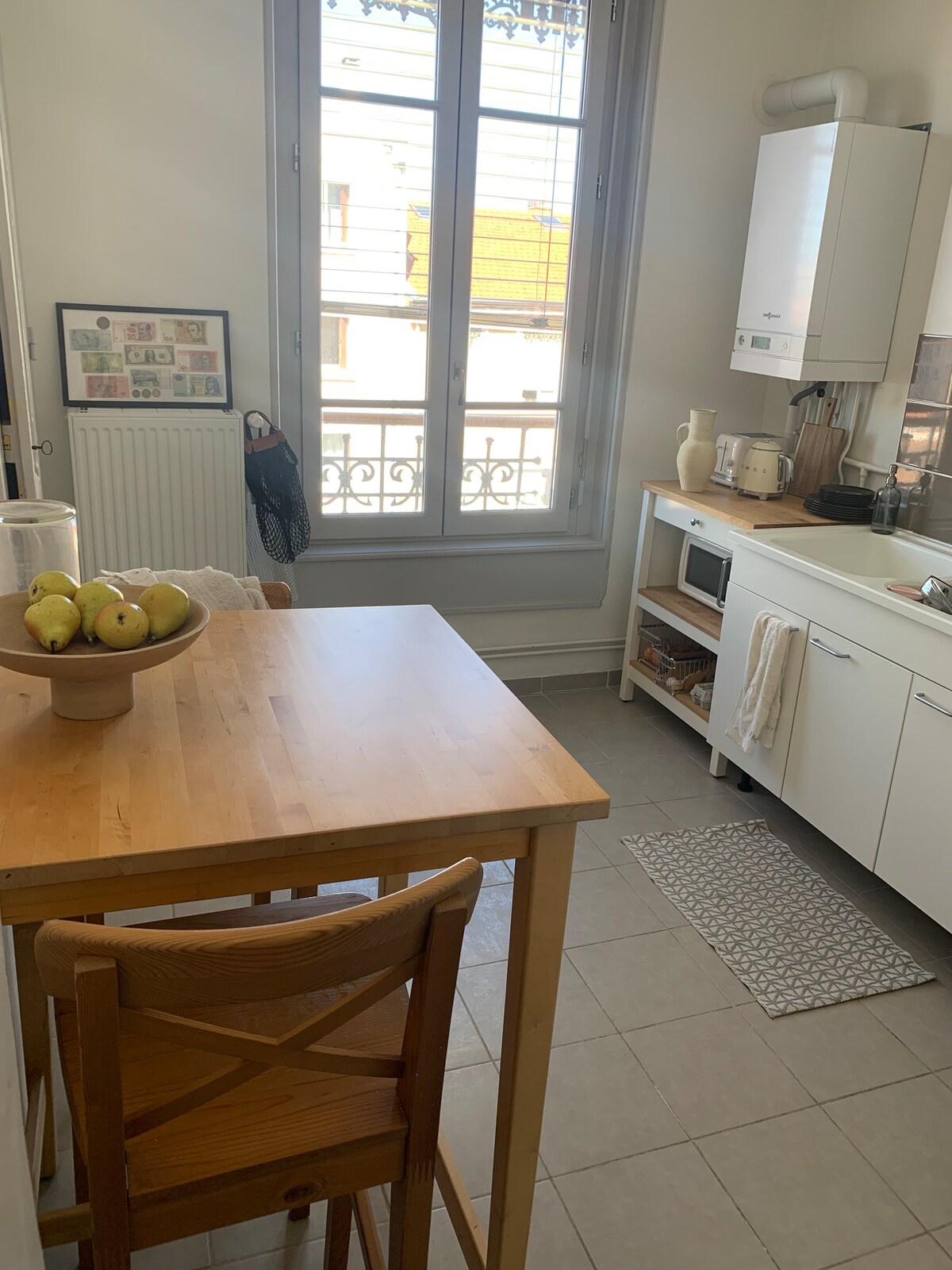 A compact kitchen space features white cabinetry and modern appliances. A small wooden table holds a bowl of green apples, positioned near a window that allows natural light to illuminate the area. A dish rack and a patterned mat are also visible.