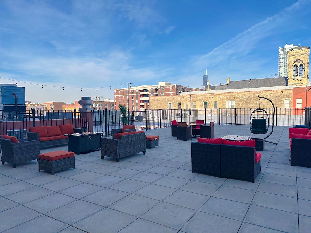A spacious rooftop terrace is displayed, featuring multiple seating arrangements with dark wicker furniture and red cushions. The area is surrounded by railings and offers a view of urban buildings under a clear blue sky.