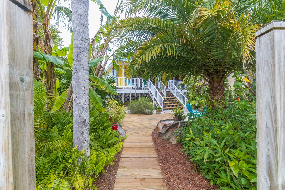 A wooden pathway is flanked by lush tropical foliage, including tall palms and vibrant green plants. The path leads towards a raised cottage, where a set of stairs is visible. The surrounding landscape creates a sense of privacy and natural beauty.