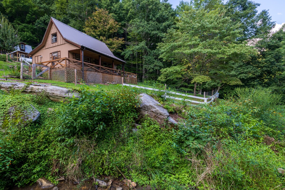 The Lee Cabin stands amidst lush greenery, showcasing a charming wooden exterior with a sloped roof. A spacious deck offers a perfect view of the surrounding trees and hillside, while the landscaping features natural stone and varied vegetation, creating a serene outdoor atmosphere.