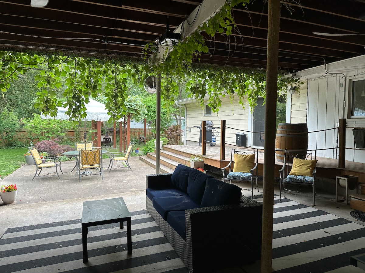The covered outdoor patio features a seating area with a dark wicker couch and coffee table, set against a striped rug. Surrounding plants create a natural canopy, and a few yellow and blue cushions add color. The yard is visible, showcasing greenery and outdoor seating.