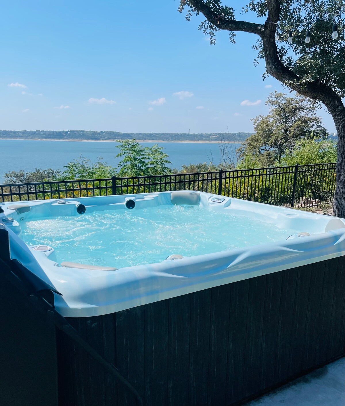 A spacious hot tub is positioned outdoors, offering inviting views of the lake in the background. The surrounding area features a black fence and lush greenery, enhancing the relaxing environment. Bright blue skies and soft clouds complement the serene landscape.
