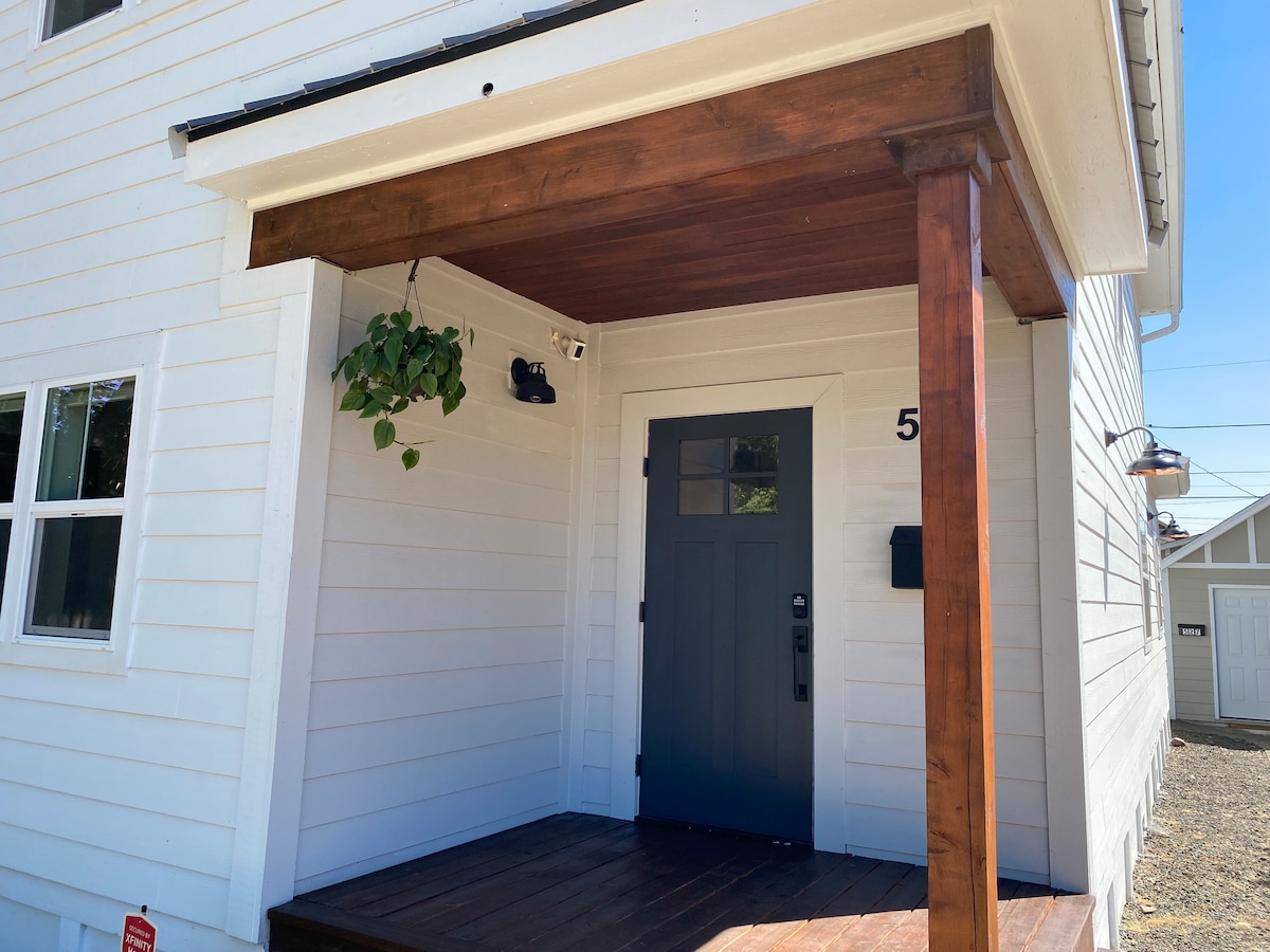 A welcoming entrance features a covered porch with a wooden beam and a hanging plant. The front door is painted a dark blue, and house numbers are displayed prominently. Surrounding windows allow natural light to illuminate the entryway's bright white siding.