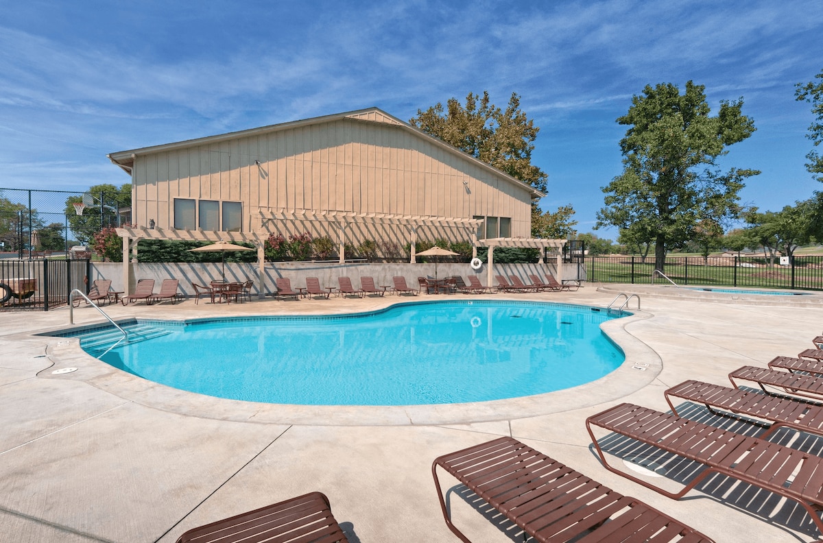 A seasonal outdoor pool is surrounded by sun loungers, offering a spacious area for relaxation. Lush greenery and a building with large windows can be seen in the background, creating an inviting environment for guests.