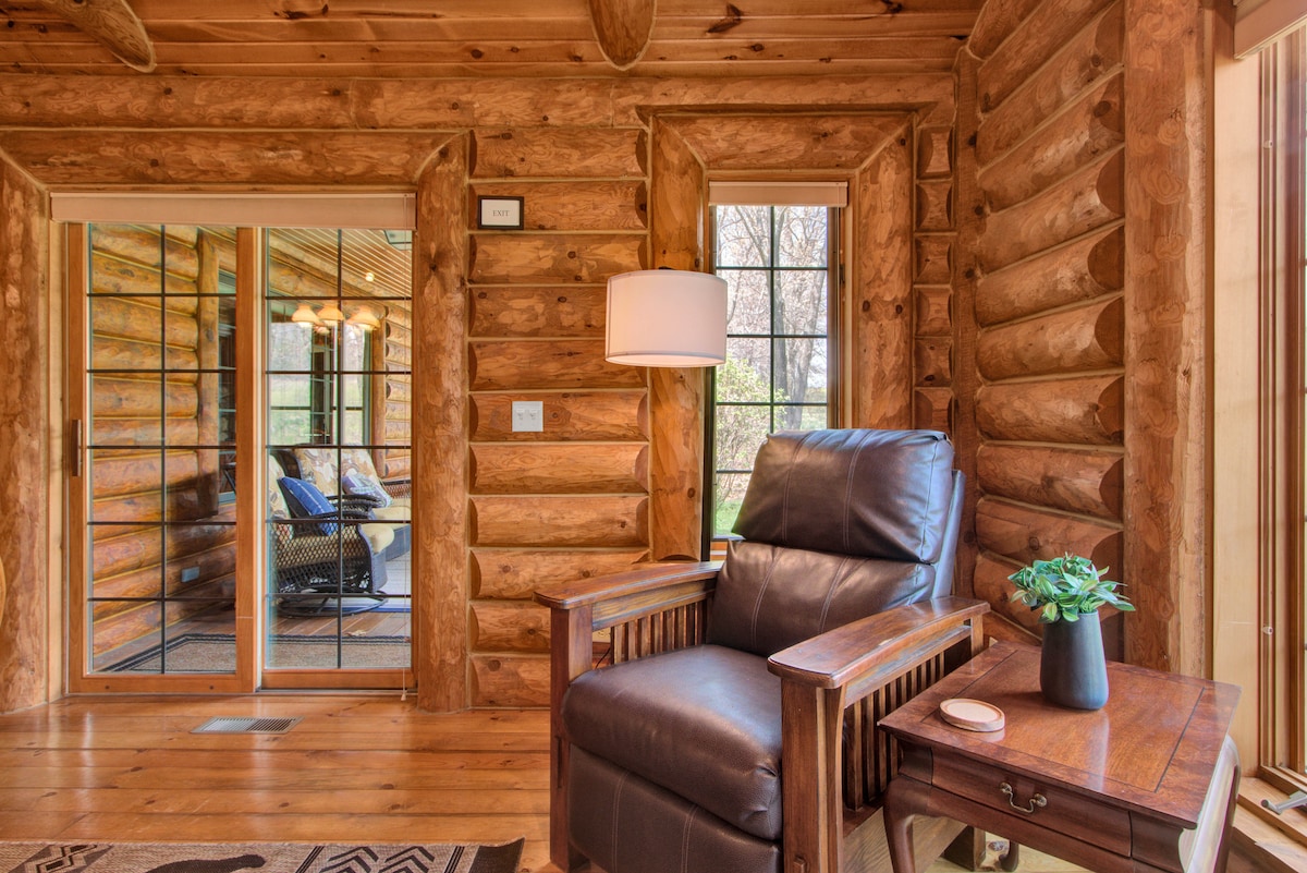 A cozy seating area is presented with a leather chair beside a small wooden table. Natural light filters through large glass doors that lead to an outdoor space. The warm log walls create an inviting ambiance, and a small plant enhances the tranquil setting.