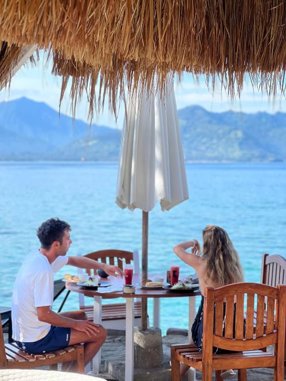 A table set for two is positioned under a thatched roof, offering views of the ocean and distant mountains. Drinks and food are placed on the table, while an umbrella provides shade. The tranquil setting highlights the inviting outdoor dining experience.