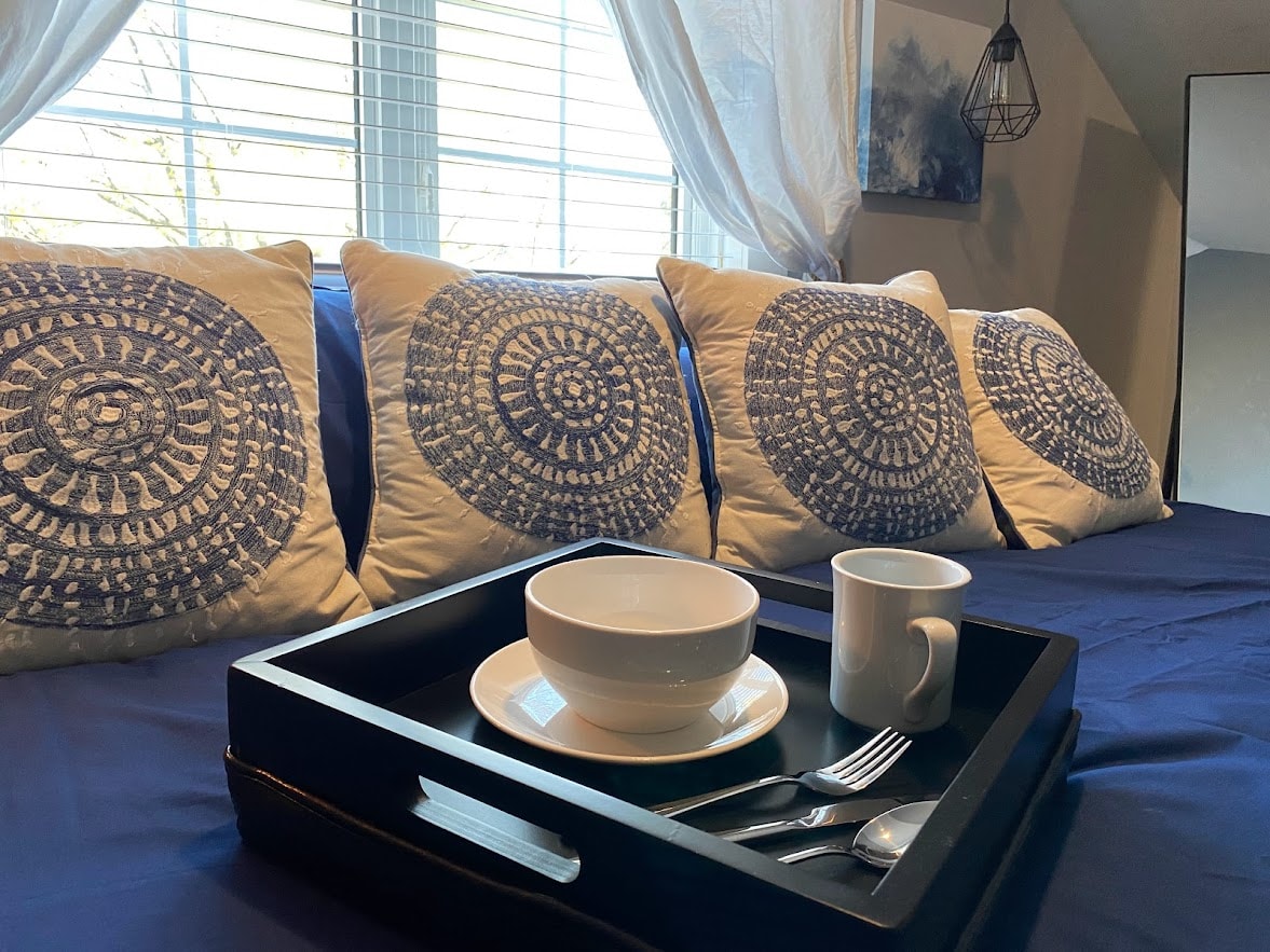 A cozy bed is adorned with decorative pillows featuring a circular design. A serving tray holds a bowl, cup, and utensils, positioned on the bedspread. Natural light filters through the window, illuminating the space.