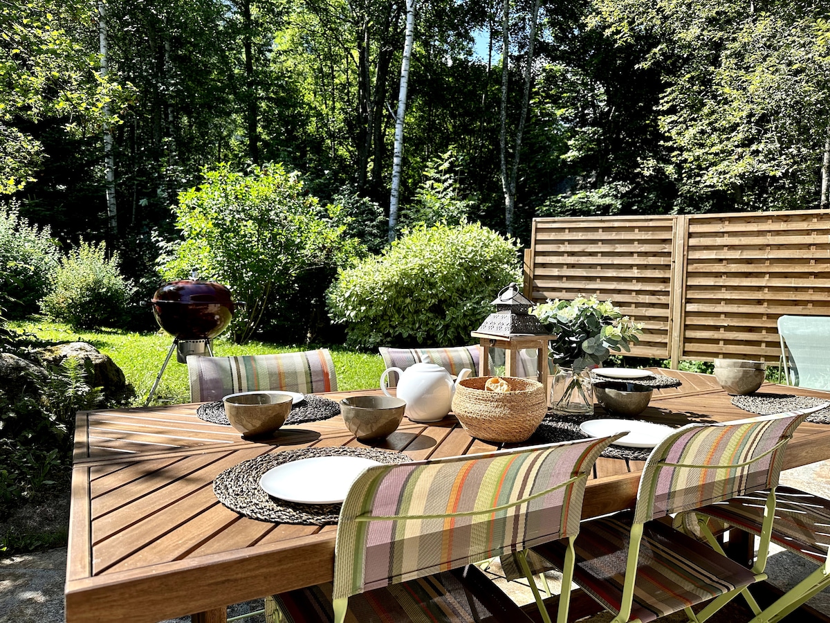 A wooden dining table is set for outdoor meals, featuring plates, bowls, and decorative items. Colorful striped chairs surround the table, while a charcoal grill and lush greenery provide a tranquil backdrop. A wooden fence creates privacy around the seating area.