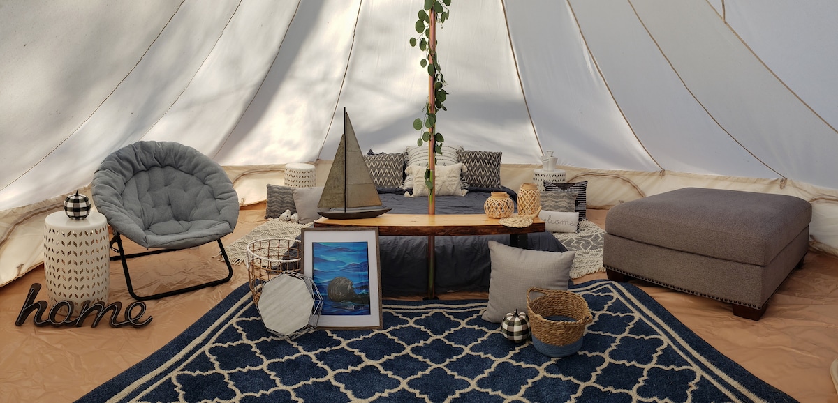 A spacious glamping tent interior is highlighted by a large, comfortable seating area furnished with a plush chair, neutral-toned cushions, and a low wooden table. A decorative sailboat and framed artwork are showcased on the floor. A soft area rug adds warmth to the natural tones.