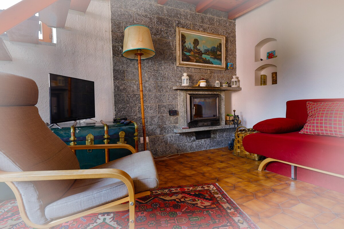 A cozy living area features a red sofa and a comfortable armchair positioned beside a stone fireplace. A small television is placed on a wooden cabinet. Decorative items are displayed on built-in shelves, and a vibrant area rug adds warmth to the space.