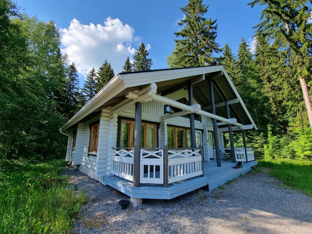 A two-story log villa features a spacious porch with wooden railings and several large windows allowing ample natural light. Surrounding greenery includes tall trees, enhancing the serene environment. The exterior is painted in light colors, contrasting with the vibrant landscape.
