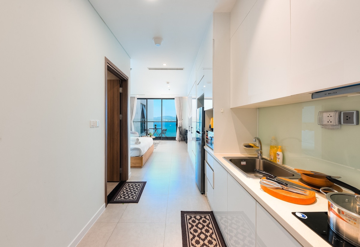 The kitchen area features modern appliances and ample counter space. Natural light illuminates the open layout, showcasing a seamless transition to the living space with a view of the sea in the background. The entrance leads to a spacious bedroom area with a large bed.