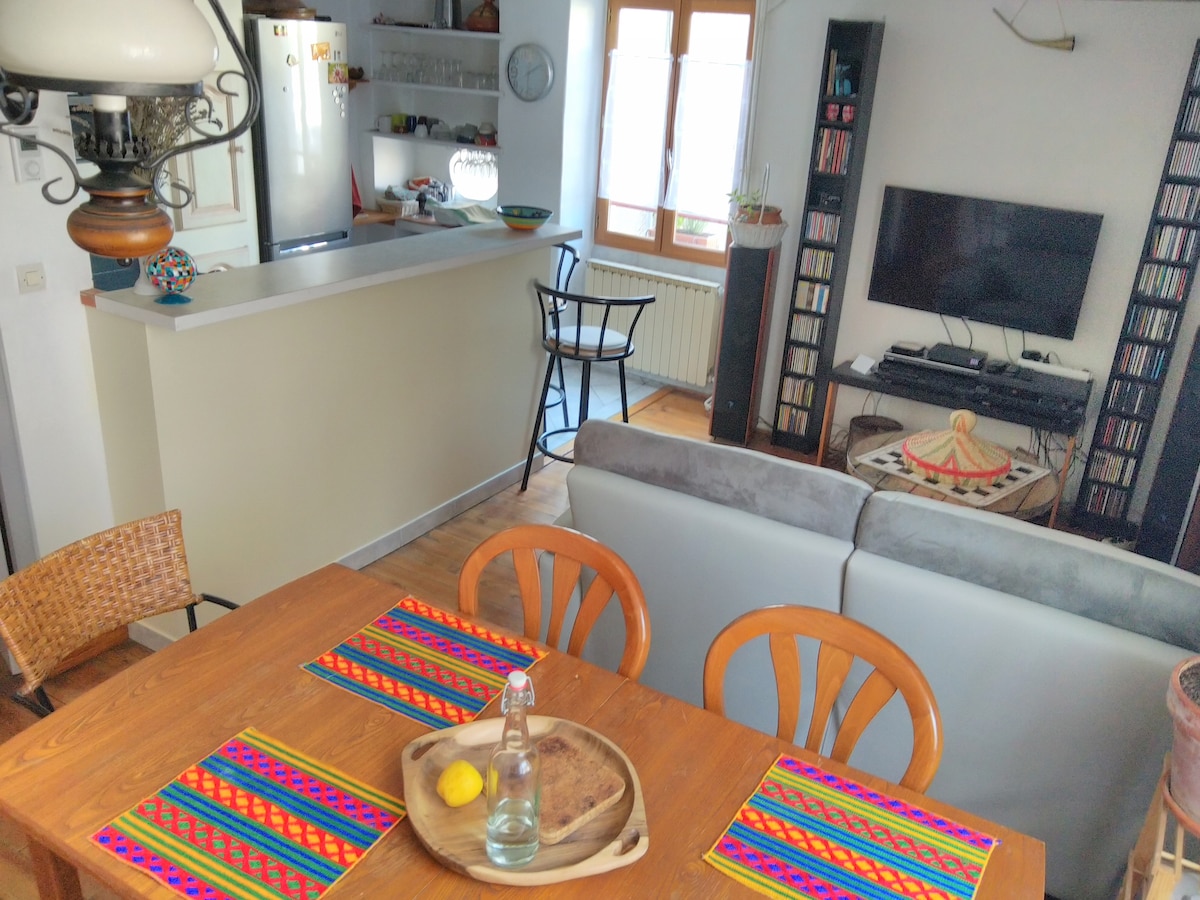 A dining table with colorful placemats is set for meals, accompanied by wooden chairs. A cozy living area features a gray sofa facing a mounted TV and a bookshelf filled with DVDs. A well-equipped kitchen area is visible in the background.
