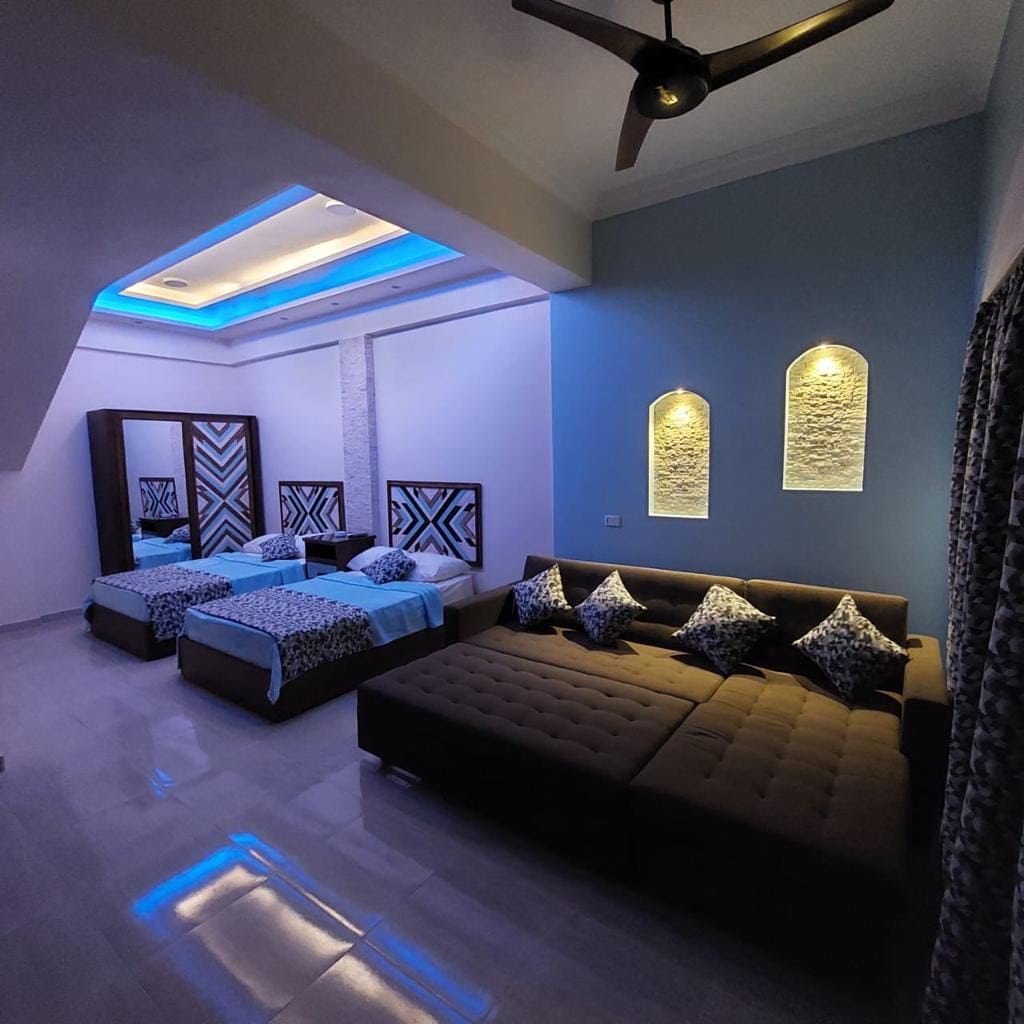 A spacious studio is highlighted by a blue-accented ceiling and soft overhead lighting. The room features two beds with coordinating blue linens and a large sectional sofa. Decorative wall niches add visual interest, while patterned curtains frame a large window.