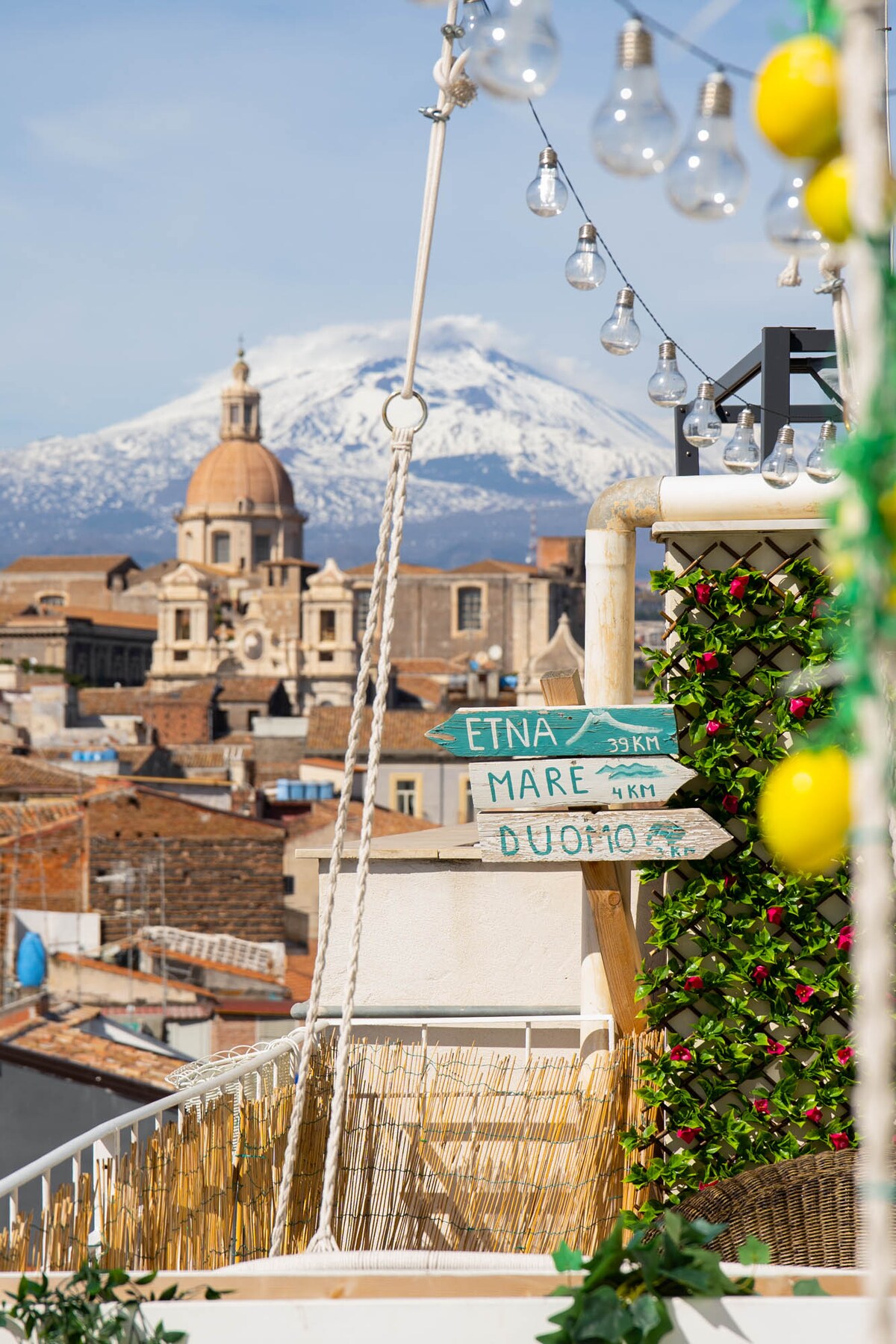 A vibrant rooftop terrace is depicted, featuring decorative string lights and lush greenery. Directional signs point to local landmarks including Etna and the Duomo. The backdrop displays the snow-capped summit of Mount Etna, alongside the charming rooftops of the historic city.