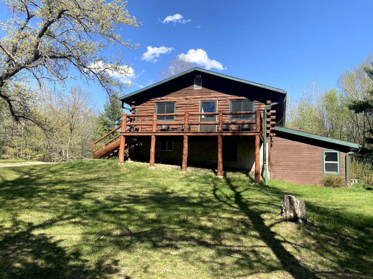 The exterior of a rustic log cabin is visible, featuring a sloped roof and large wooden deck with railings. The cabin is surrounded by a grassy area, with trees and blue sky in the background, creating a serene countryside setting.