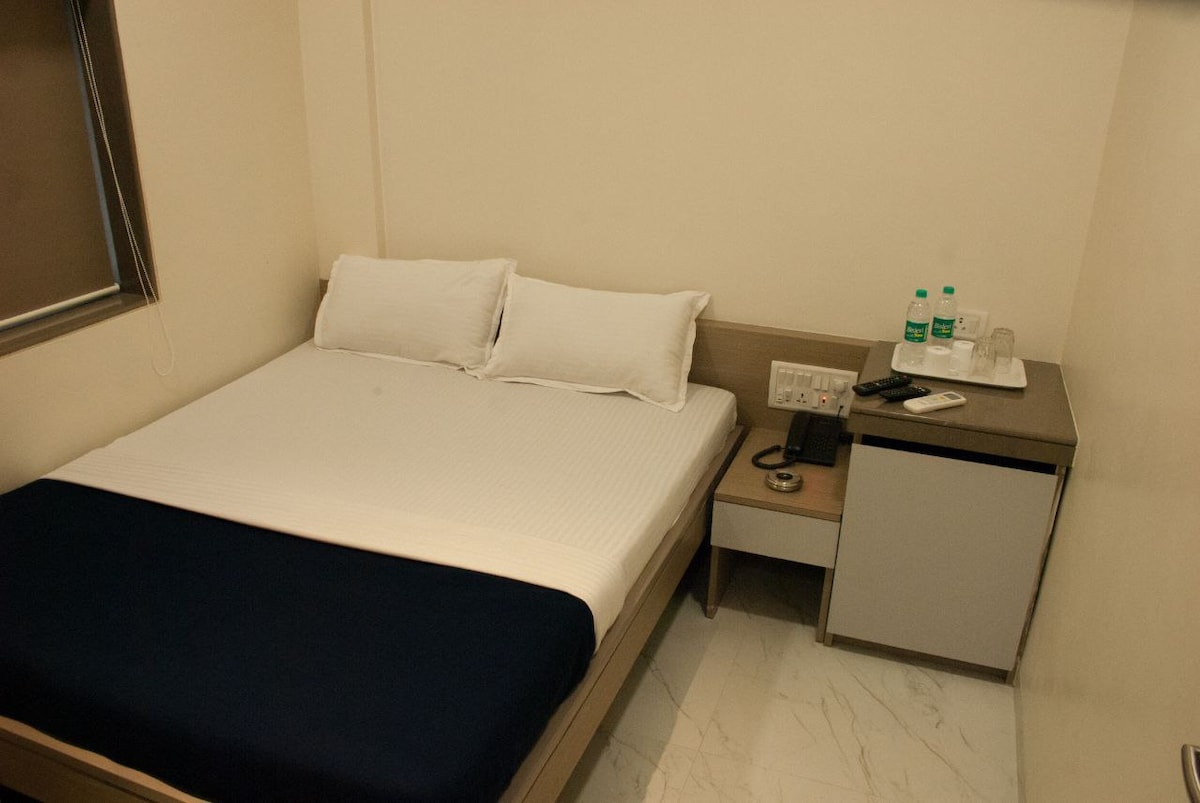A serene sleeping area is presented with a spacious bed dressed in crisp white linens and a navy blue throw. A modern bedside table holds a telephone and two bottles of water, while a small fridge is positioned beside it. Natural light enters through the nearby window.