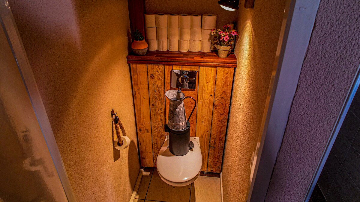 A compact bathroom features a modern toilet positioned against a wooden backdrop. Above the toilet, a series of potted plants and decorative items add a natural touch. Soft lighting illuminates the space, creating a calm atmosphere.