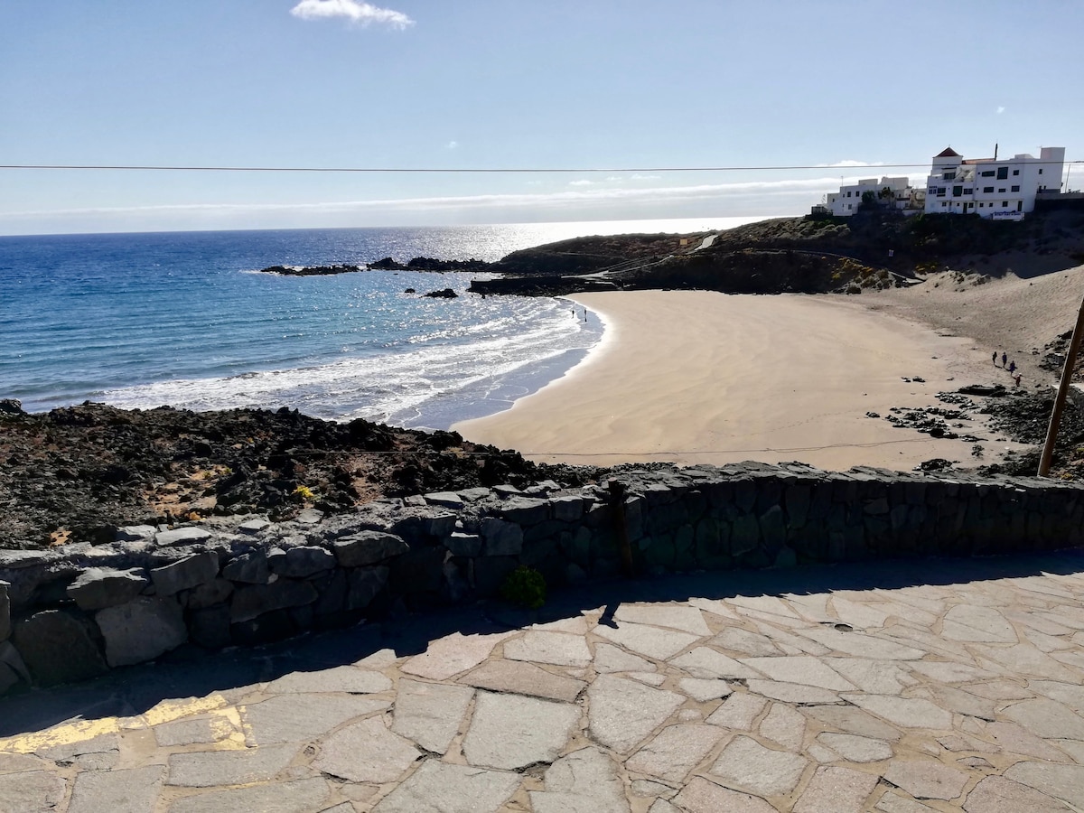 A serene coastal view showcases a sandy beach bordered by gentle waves. Rocky outcrops frame the shoreline, while distant white buildings dot the landscape. Sunlight sparkles on the water, creating a peaceful ambiance, ideal for relaxation and outdoor activities.