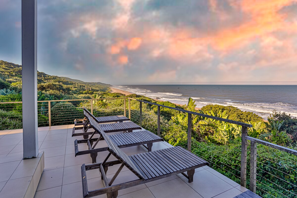 A spacious deck is equipped with wooden lounge chairs, offering views of the lush green landscape and vast ocean. The sky reflects warm hues during sunset, creating a serene atmosphere for relaxation.