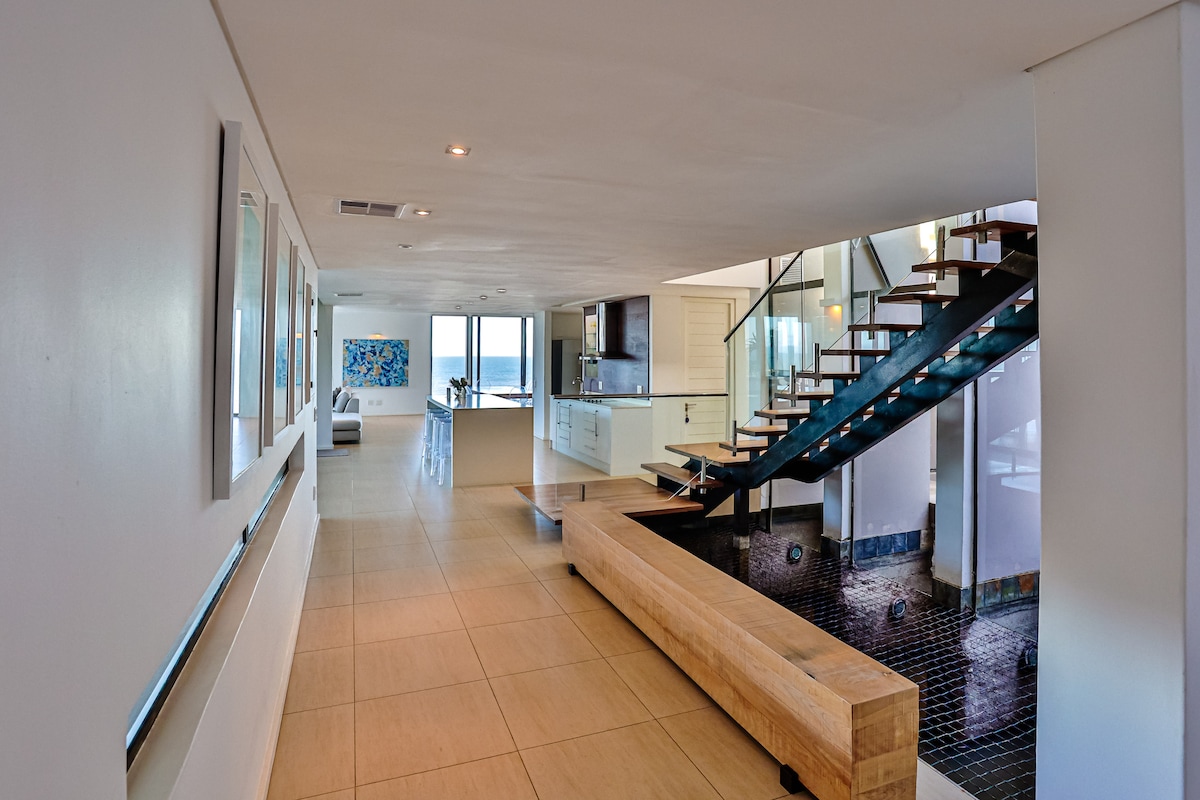 An open and airy entrance hall features a sleek staircase leading to the upper floor. A modern kitchen is visible in the background, with clear views of the large windows framing the ocean beyond. A wooden bench separates the hallway from the Koi pond below.