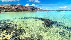 A serene coastal scene captures clear turquoise water gently lapping against rocky shorelines. Shallow areas reveal a sandy bottom and scattered rocks, while distant hills provide a backdrop under a bright blue sky adorned with fluffy white clouds.