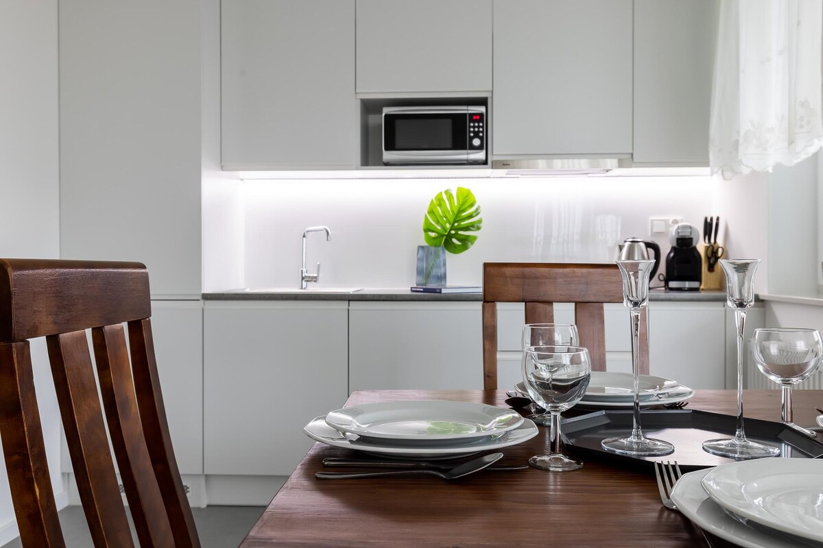 A modern kitchen area is shown, featuring a clean white design with sleek cabinetry and a countertop. A dining table is set with elegant glassware and plates. The kitchen is equipped with a microwave and a decorative plant adds a touch of greenery.