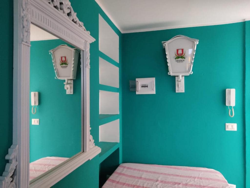 A vibrant bedroom features refreshing turquoise walls and a spacious bed adorned with a striped blanket. A large, decorative mirror reflects the room's design, while framed artwork and an intercom add functional elegance to the space.