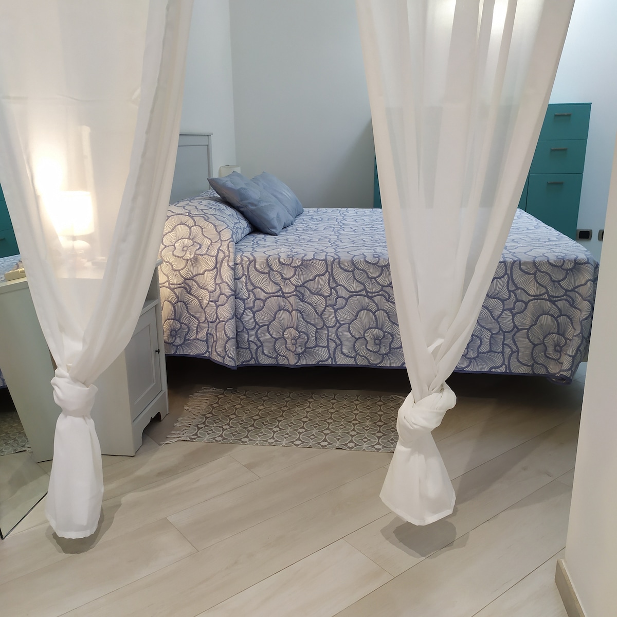 A spacious bedroom features a neatly made bed with a floral-patterned coverlet and soft blue pillows. Flowing white curtains create a serene ambiance, while a decorative rug and subtle bedside lamps add to the inviting decor. Light filters gently through the airy space.
