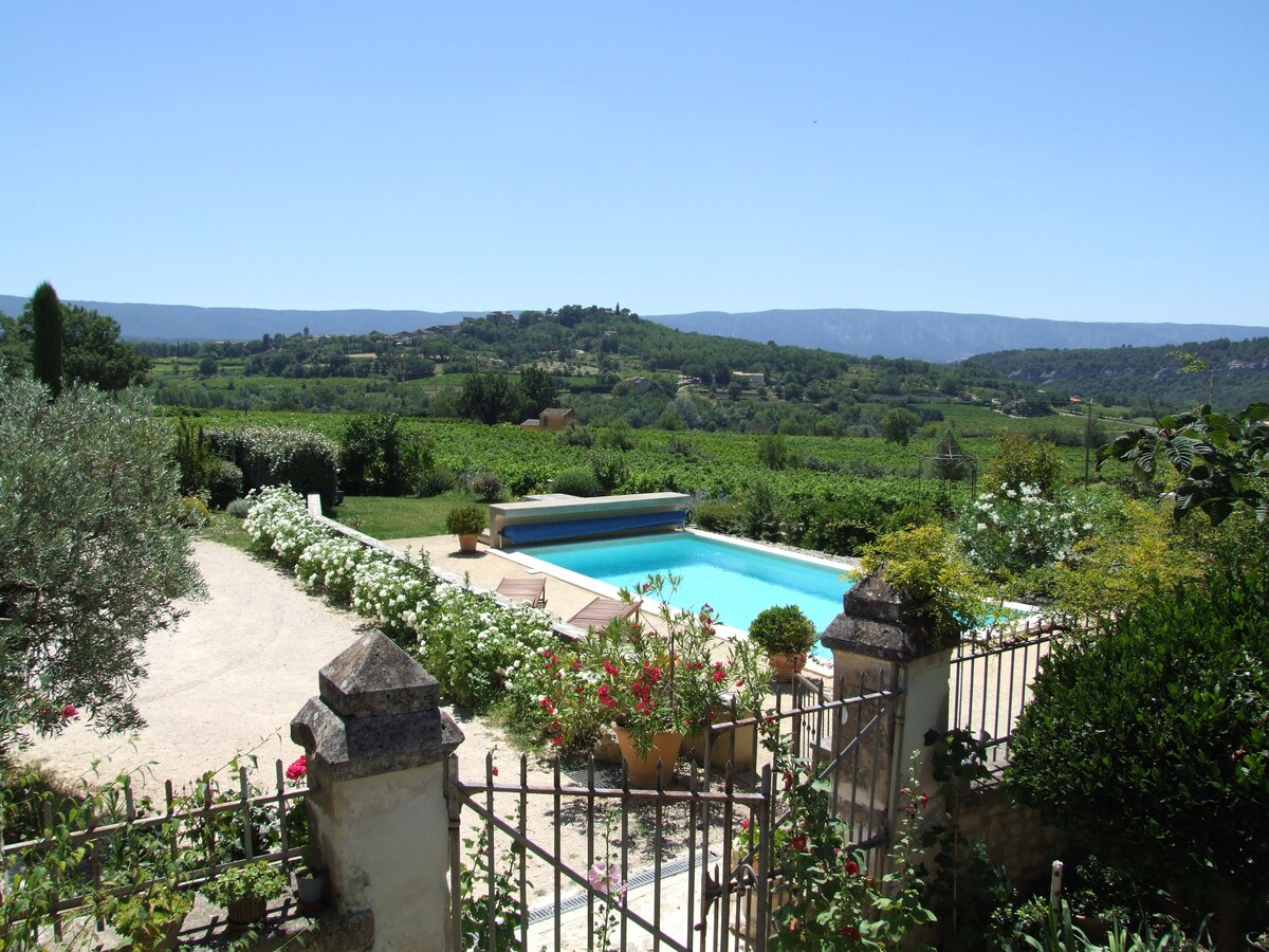 A serene outdoor landscape features a swimming pool surrounded by lush gardens and blooming flowers. Rolling hills and vineyards are visible in the background under a clear blue sky, creating a peaceful atmosphere. Stone fencing outlines the property, blending naturally with the surroundings.