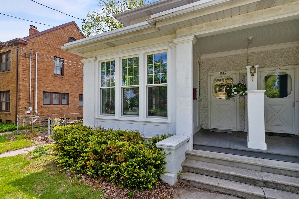 The exterior entrance features a set of double doors, framed by large windows that allow natural light to fill the entryway. Lush green landscaping with manicured bushes enhances the welcoming atmosphere of the property. A set of stone steps leads up to the porch.