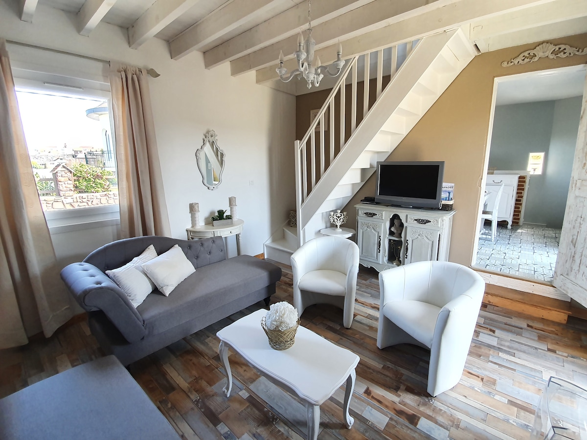 The living room features a light-colored staircase leading to the upper level, with a gray sofa and two white armchairs arranged around a small coffee table. Natural light floods the room from a window, highlighting wooden flooring and classic decor elements.