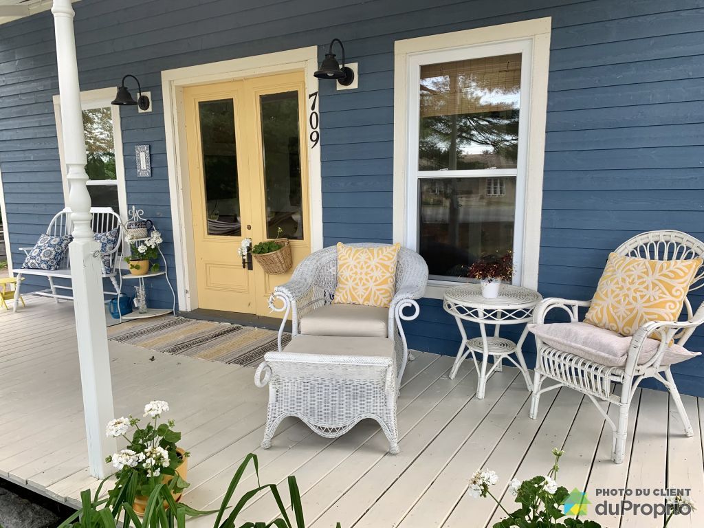 A front porch features white wicker chairs adorned with yellow cushions and a small table. The blue exterior of the house enhances the inviting atmosphere, complemented by flowers in the foreground and large windows allowing natural light.