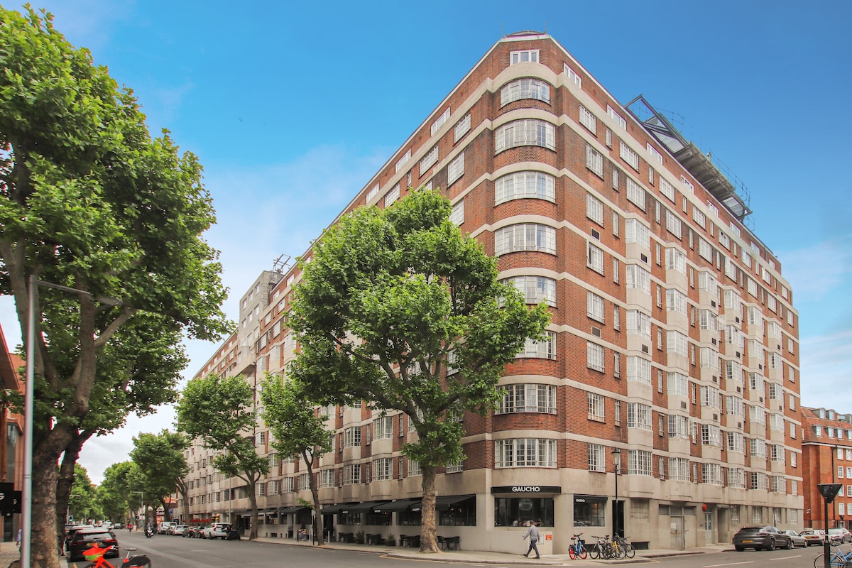 A modern brick building with a distinct architectural style is viewed from the street. Large windows punctuate the facade, while leafy trees line the sidewalk, enhancing the urban atmosphere. Balconies can be seen along the upper levels, adding to the building's contemporary appeal.