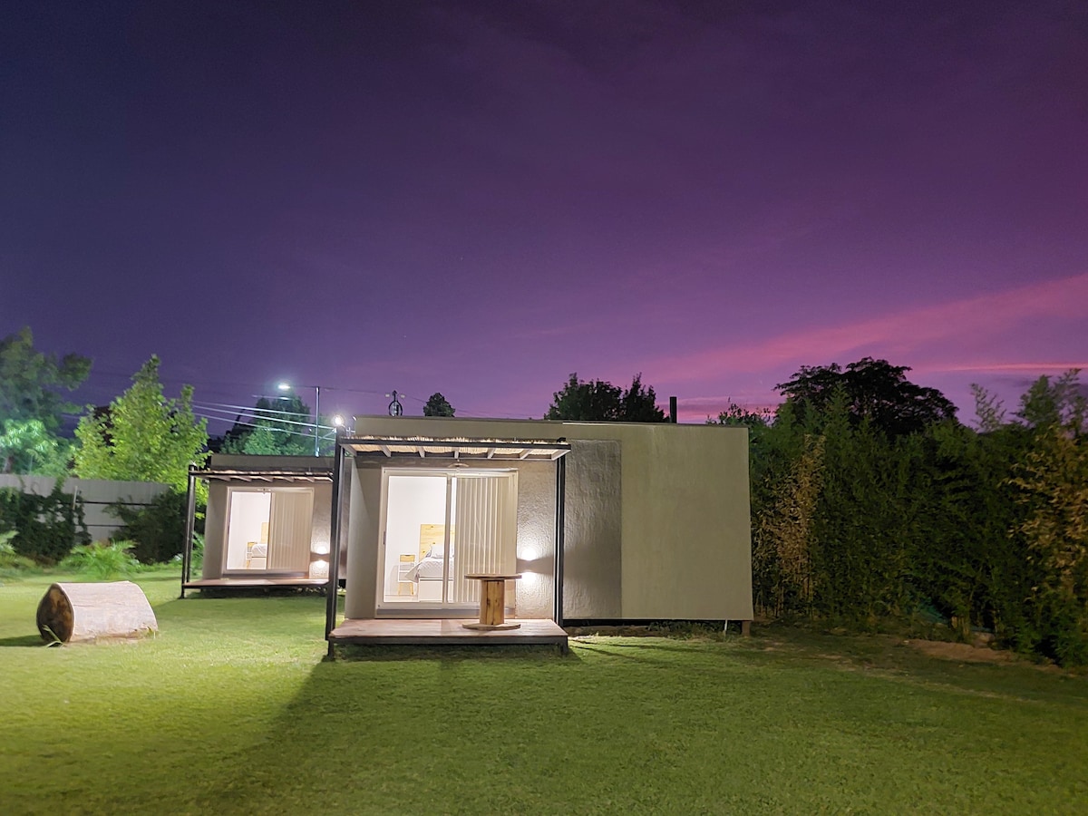 A modern structure is set in a lush green yard, illuminated under a twilight sky filled with soft purple hues. Outdoor seating is available on a wooden deck, providing a view of the serene surroundings, including trees and distant lights.