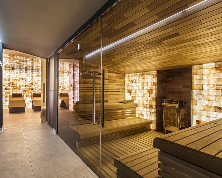 The wellness area features a modern sauna with wooden benches and a stone wall. Adjacent to the sauna, a relaxation area is highlighted by walls made of illuminated salt bricks and lounge chairs, creating a serene environment for guests.
