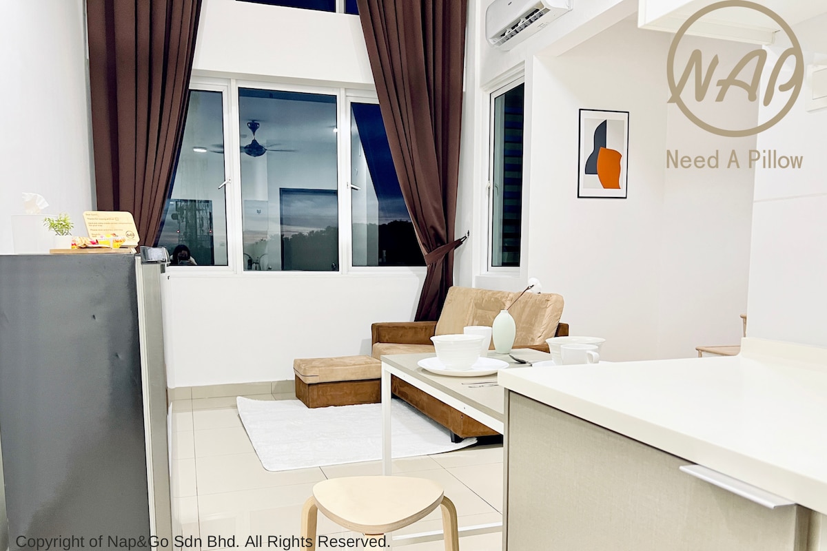 A cozy living space features a brown sofa and a wooden coffee table, accompanied by a light rug. Large windows with dark curtains provide natural light, while decorative items and a small kitchen area enhance the functionality of the room.