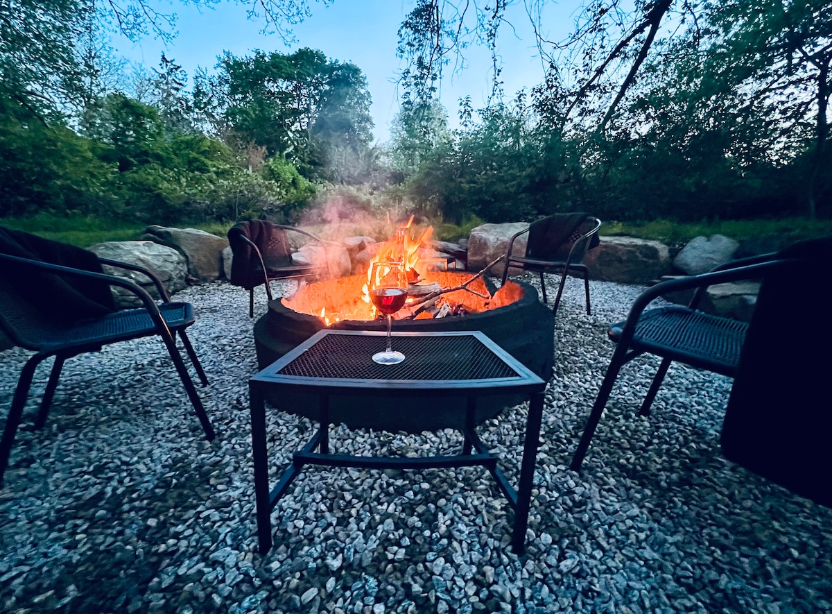A fire pit surrounded by a gravel area is shown, with four black metal chairs positioned around it. A glass of red wine rests on a dark table, while smoke gently rises from the flickering flames, creating a cozy ambiance among the trees.